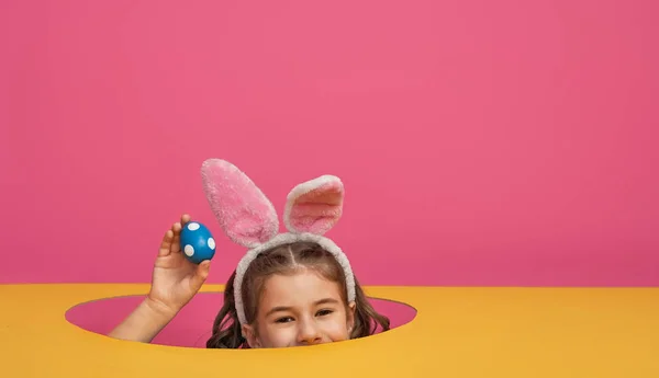 Cute little child wearing bunny ears on Easter day. Girl with painted eggs on bright pink and yellow background.