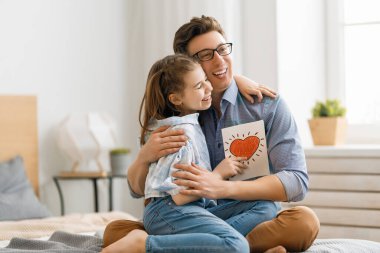 Babalar günün kutlu olsun! Çocuk kızı babasını tebrik ediyor ve ona kartpostal veriyor. Baba ve kız gülümsüyor ve sarılıyorlar. Aile tatili ve birliktelik.