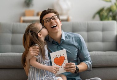 Babalar günün kutlu olsun! Çocuk kızı babasını tebrik ediyor ve ona kartpostal veriyor. Baba ve kız gülümsüyor ve sarılıyorlar. Aile tatili ve birliktelik.