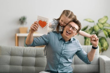 Babalar günün kutlu olsun! Çocuk kızı babasını tebrik ediyor ve ona kartpostal veriyor. Baba ve kız gülümsüyor ve sarılıyorlar. Aile tatili ve birliktelik.