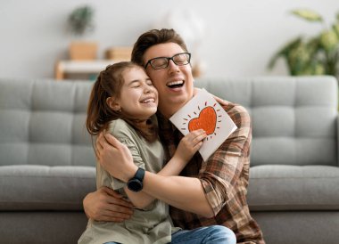 Babalar günün kutlu olsun! Çocuk kızı babasını tebrik ediyor ve ona kartpostal veriyor. Baba ve kız gülümsüyor ve sarılıyorlar. Aile tatili ve birliktelik.