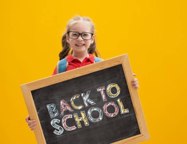 Okula dön ve mutlu ol! Şirin, çalışkan bir çocuk duvar kağıdının arka planında karatahta tutuyor. Sırt çantalı çocuk. Kız çalışmaya hazır..