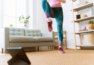 Aktif giyimli genç bir kadın evde egzersiz yaparken tabletten online kurslar izliyor..