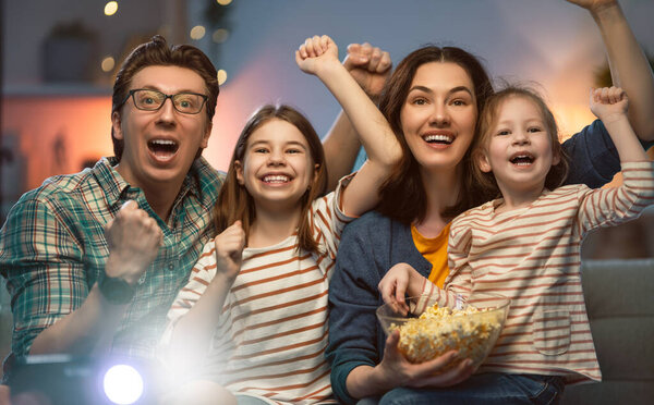 Happy family watching projector, TV, movies with popcorn in the evening at home. Mother, father and daughters spending time together.