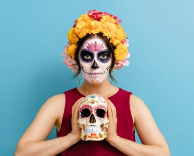 Mavi arka planda çiçek çelenkleriyle poz veren sevimli zombi. Cadılar Bayramı yaratıcı makyajlı mutlu kadın. Meksika Ölüler Günü 'nü kutlayan kız.