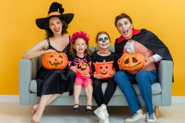 Anne, baba ve çocukları partide eğleniyorlar. Cadılar Bayramı 'nı kutlayan mutlu aile. Sarı arka planda karnaval kostümleri ve makyaj yapan insanlar var..