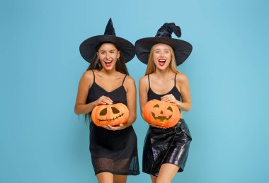 Mutlu Cadılar Bayramı! Mavi arka planda balkabakları olan iki siyah cadı kostümlü genç kadın..