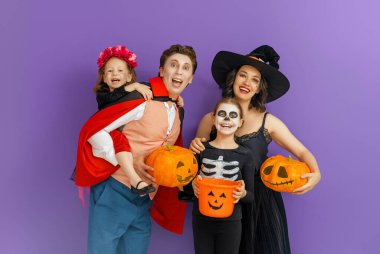 Anne, baba ve çocukları partide eğleniyorlar. Cadılar Bayramı 'nı kutlayan mutlu aile. İnsanlar mor arka planda karnaval kostümü ve makyaj giyiyorlar..