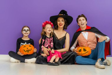 Anne, baba ve çocukları partide eğleniyorlar. Cadılar Bayramı 'nı kutlayan mutlu aile. İnsanlar mor arka planda karnaval kostümü ve makyaj giyiyorlar..