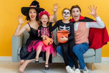Anne, baba ve çocukları partide eğleniyorlar. Cadılar Bayramı 'nı kutlayan mutlu aile. Sarı arka planda karnaval kostümleri ve makyaj yapan insanlar var..