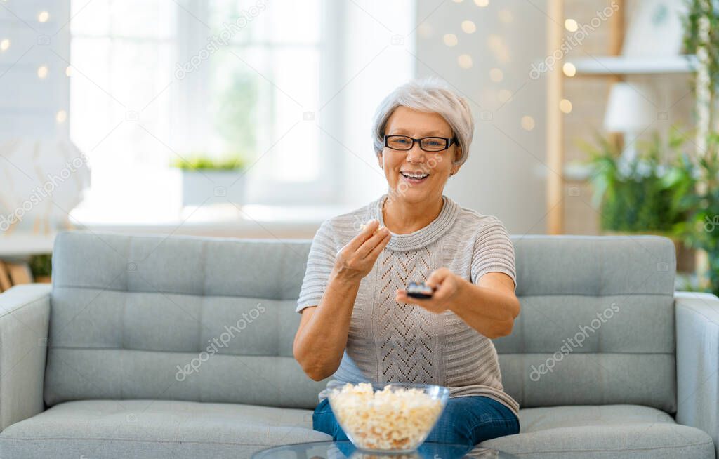 Senior woman watching projector, TV, movies with popcorn. Retiree spending time at home.