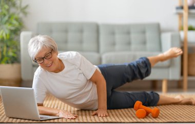Aktif giyimde kıdemli bir kadın evde egzersiz yaparken dizüstü bilgisayarda ders izliyor..