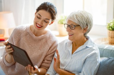 Genç ve yaşlı kadınlar tablet bilgisayar kullanıyorlar. Kız, anneye evde kalarak eğlenmesi için yardım ediyor..