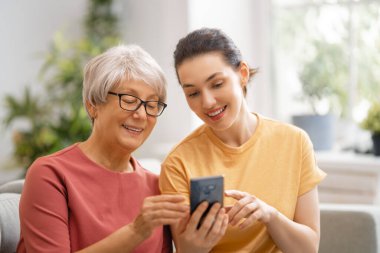 Genç ve yaşlı kadınlar akıllı telefon kullanıyor. Kız, anneye evde kalarak eğlenmesi için yardım ediyor..