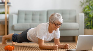 Aktif giyimde kıdemli bir kadın evde egzersiz yaparken dizüstü bilgisayarda ders izliyor..