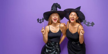 Mutlu Cadılar Bayramı! Siyah cadı kostümlü iki genç kadın mor arka planda partide..