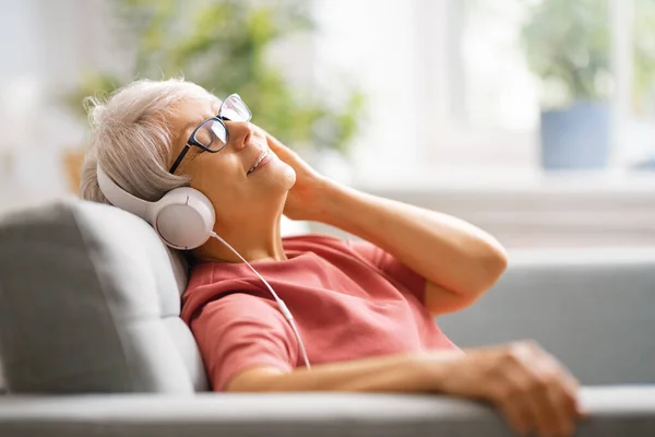 老年妇女坐在家里的沙发上 用耳机听音乐 — 图库照片