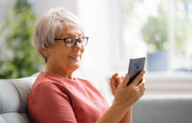 Neşeli, güzel, son sınıf öğrencisi kadın akıllı telefon kullanıyor. Evdeki kanepede oturuyor..
