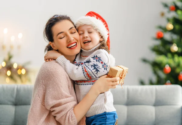 Merry Christmas and Happy Holidays! Cheerful mom and her cute daughter girl exchanging gifts. Parent and little child having fun near tree indoors. Loving family with presents in room.