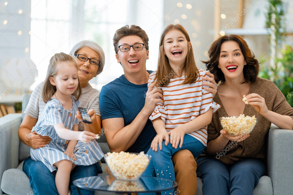 Happy family watching TV with popcorn  at home. Granny, mother, father and daughters spending time together.