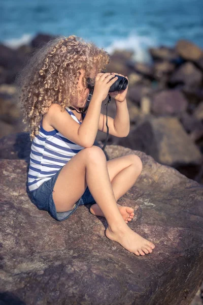 Pieni Tyttö Katselee Kauas Kiikarit Päässä Istuu Kivellä Lähellä Merta — kuvapankkivalokuva