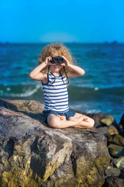 小女孩看着远处的望远镜 坐在岩石附近的海 — 图库照片