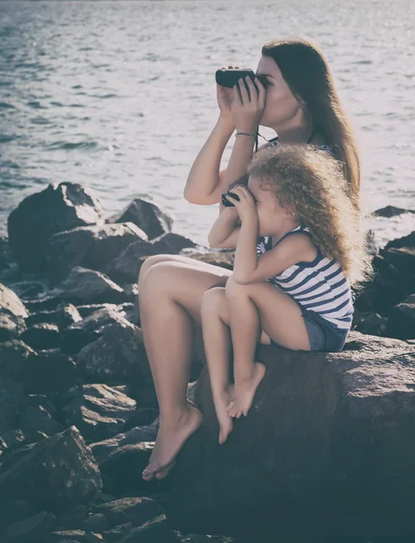 小女孩和母亲看着遥远的望远镜 坐在岩石附近的海 — 图库照片