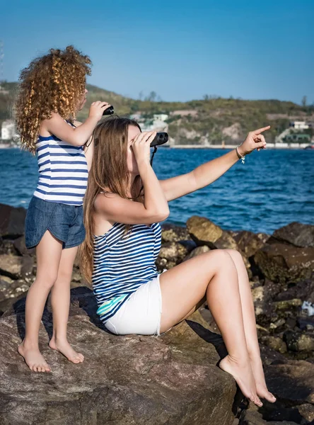 小さな女の子と母岩の上に座って 双眼鏡を使って遠くを見て近くの海 — ストック写真