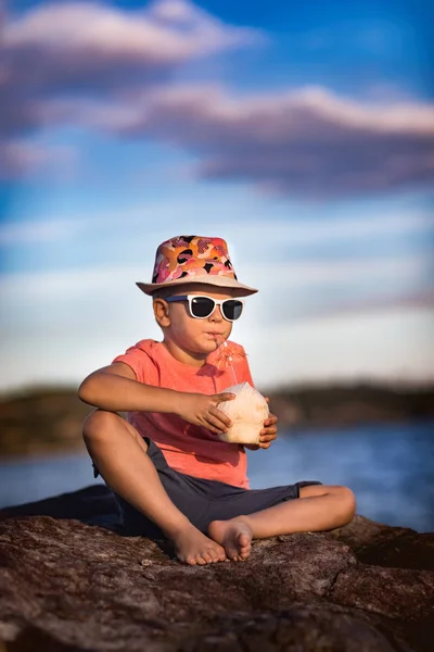 海の近くの岩の上に座ってココナッツを飲む男の子 — ストック写真