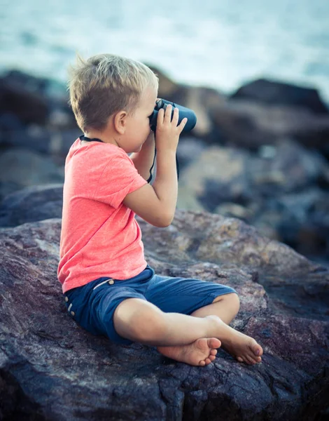 小男孩看着远处的望远镜 坐在岩石附近的海 — 图库照片