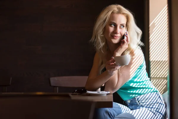 Glückliche Frau trinkt Kaffee mit Telefon am Fenster mit Jalousien. — Stockfoto
