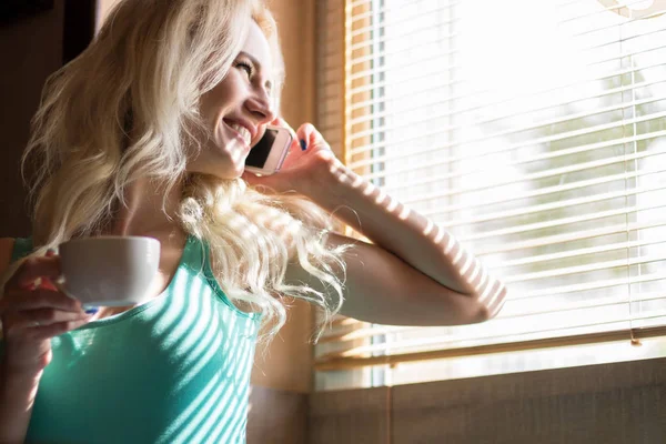 Mutlu kadın içecekler kahve ve gülümsüyor pencere panjur ile yakın:.