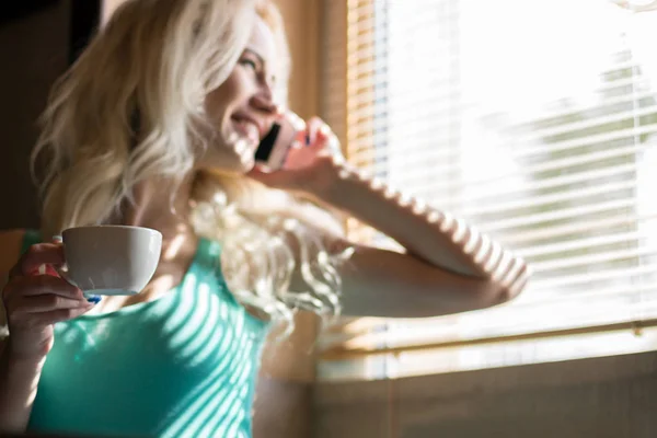 Счастливая женщина пьет кофе и улыбается у окна с жалюзи . — стоковое фото