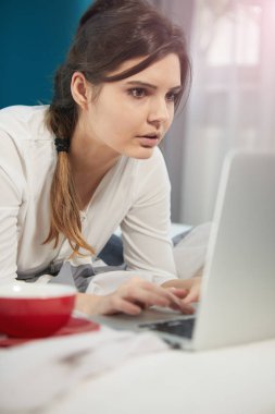 Aydınlık ve rahat yatak evde yatakta yatan dizüstü bilgisayar ile çalışan genç kadın