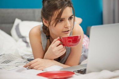 Kadın beyaz yatakta kahve yaparken dizüstü bilgisayarda tarama meşgul