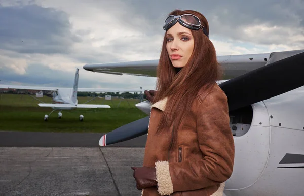 魅力的な女性パイロットを身に着けていますサングラスは彼女の小さなプライベート飛行機に対して休んで飛行場に太陽の下で立って — ストック写真