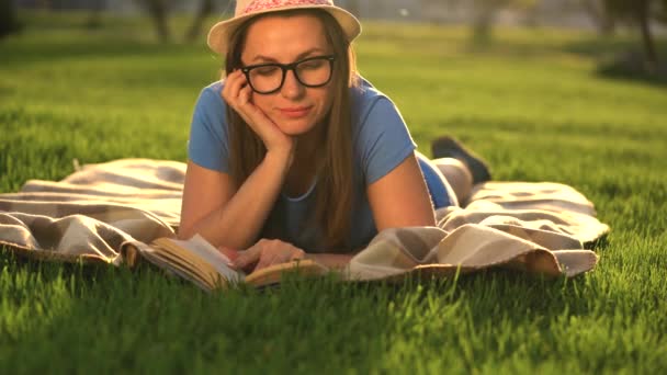 Menina de óculos lendo livro deitado em um cobertor no parque ao pôr do sol — Vídeo de Stock