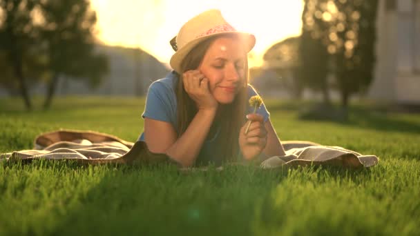 Girl with a dandelion in her hands relaxes lying down on a blanket in the park at sunset — Stock Video