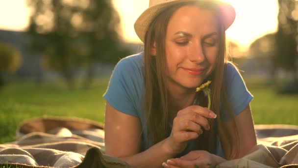 Girl with a dandelion in her hands relaxes lying down on a blanket in the park at sunset — Stock Video