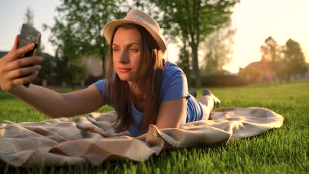 Κορίτσι κάνει μια selfie ξαπλωμένη σε μια κουβέρτα στο πάρκο στο ηλιοβασίλεμα — Αρχείο Βίντεο