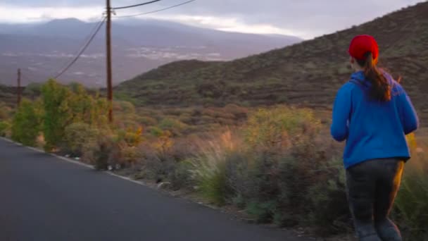 女性はビュー人けのないアスファルトの道路に沿って夕暮れ、戻って実行されます。背景に山々。スローモーション — ストック動画