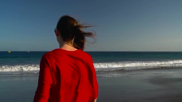 Χαρούμενος νεαρός γυναίκας με κόκκινο φόρεμα περπάτημα στο νερό στην ωκεάνια παραλία στο ηλιοβασίλεμα. Έννοια της ξέγνοιαστες σύγχρονη ζωή — Αρχείο Βίντεο
