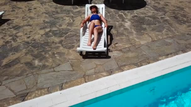 A tetején, egy nő egy kék fürdőruha sunbathes egy nyugágyon, feláll, megy a medence és úszik a medencében — Stock videók