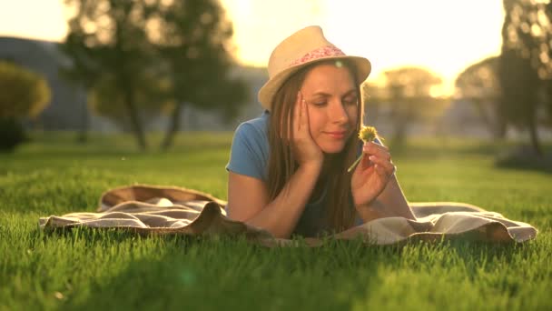 女孩用蒲公英在她的手放松躺在公园的毯子在日落 — 图库视频影像