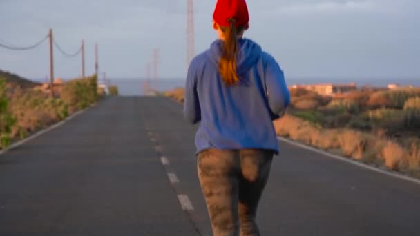 Kobieta biegnie wzdłuż drogi asfaltowej opuszczony o zachodzie słońca, powrót widok. Góry na tle. Zwolnionym tempie — Wideo stockowe