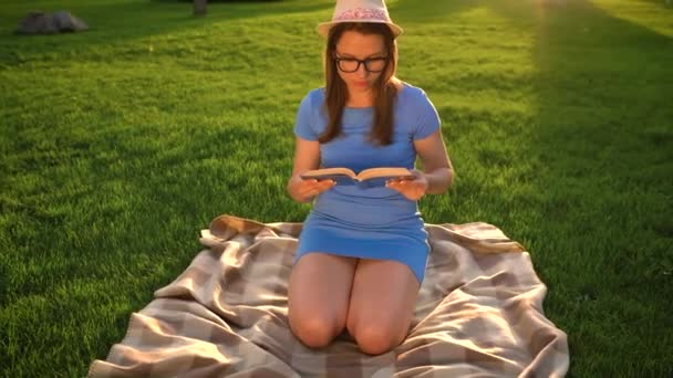 Ragazza in occhiali lettura libro seduto su una coperta nel parco al tramonto — Video Stock