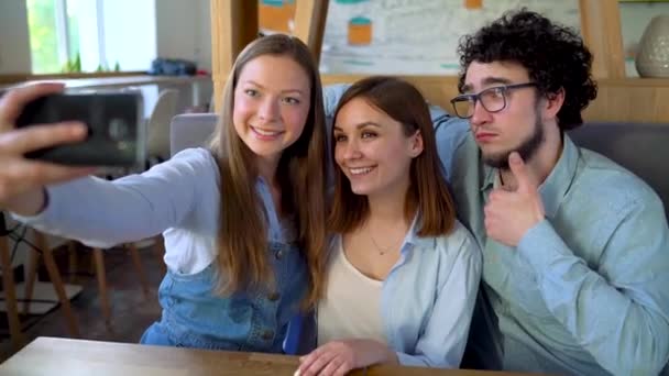 Três amigos se sentam em um café, se divertem se comunicando e fazem selfie — Vídeo de Stock