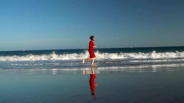 Jovem feliz em vestido vermelho corre ao longo da praia do oceano ao pôr do sol. Conceito de vida moderna despreocupada. Movimento lento — Vídeo de Stock