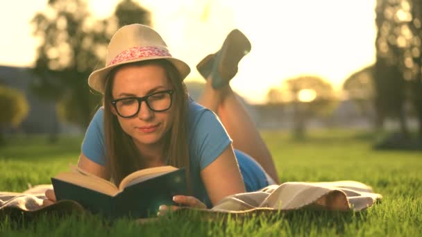 Mädchen mit Brille liest Buch und legt sich bei Sonnenuntergang auf eine Decke im Park — Stockvideo