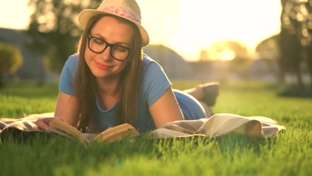 Girl in glasses reading book lying down on a blanket in the park at sunset — Stock Video